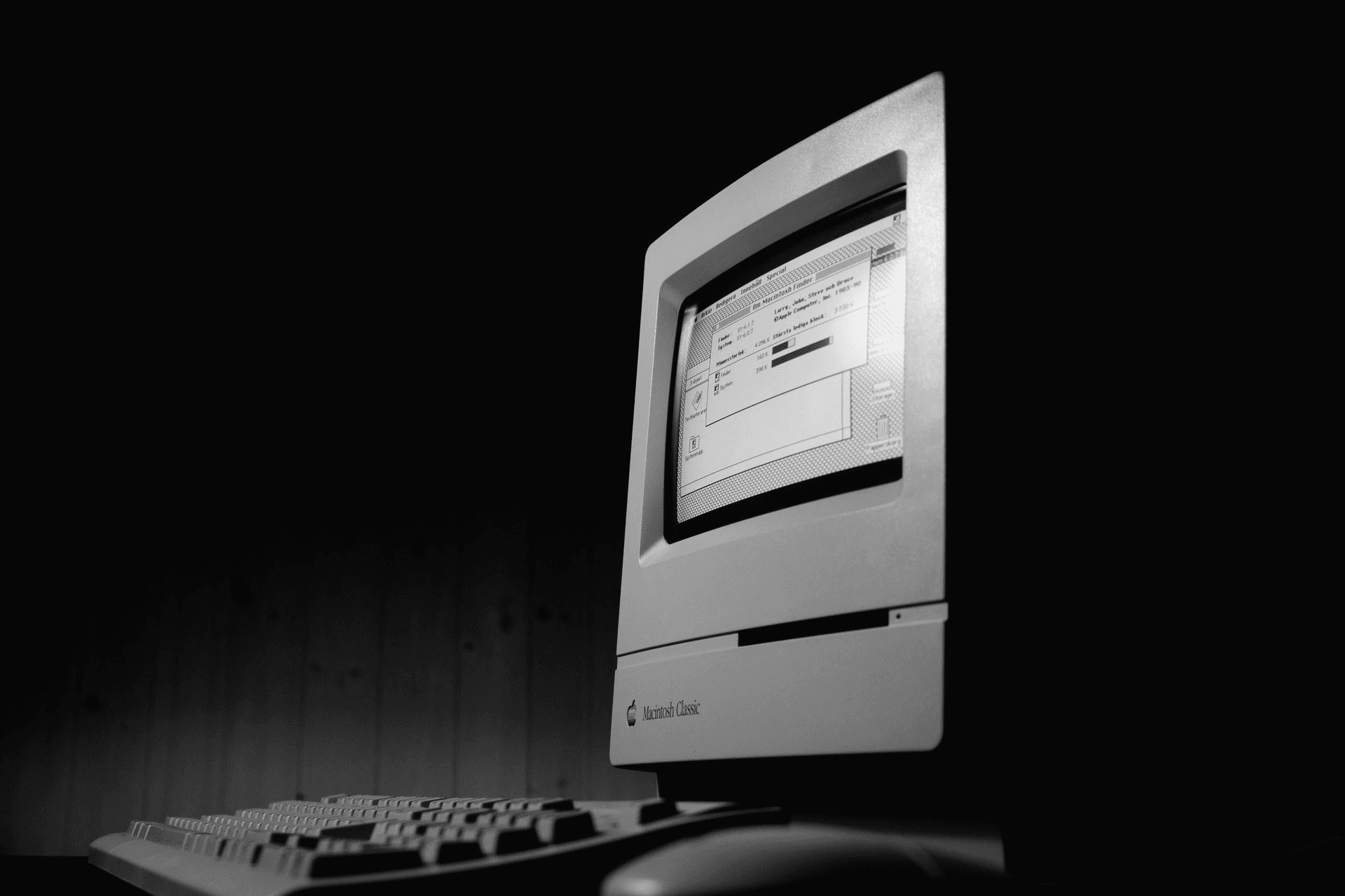 A vintage Macintosh Classic computer glowing in a dark room, with its keyboard and mouse in the foreground.